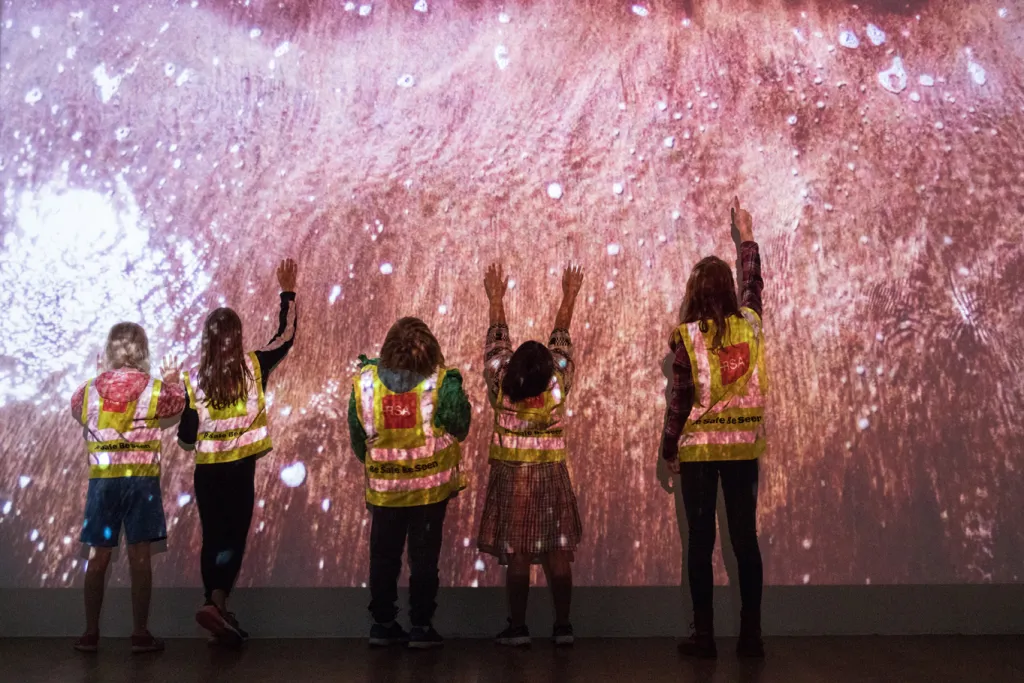 Five children in high-visability jackets facing a wall with a projected video of dusty pinkish tones.