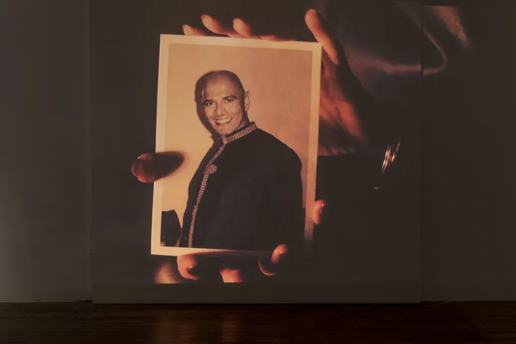 A large projection on a white wall in a dark room, the video depicts a pair of hands holding up a black and white image of a man.