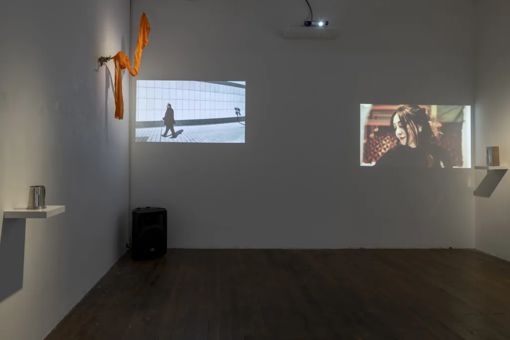 Exhibition documentation of a white wall gallery with a wooden floor, two small projections depicting a close of a women and a person walking, a sculpture of orange fabric hangs on the left wall and small white shelf with a silver jug sits below it.