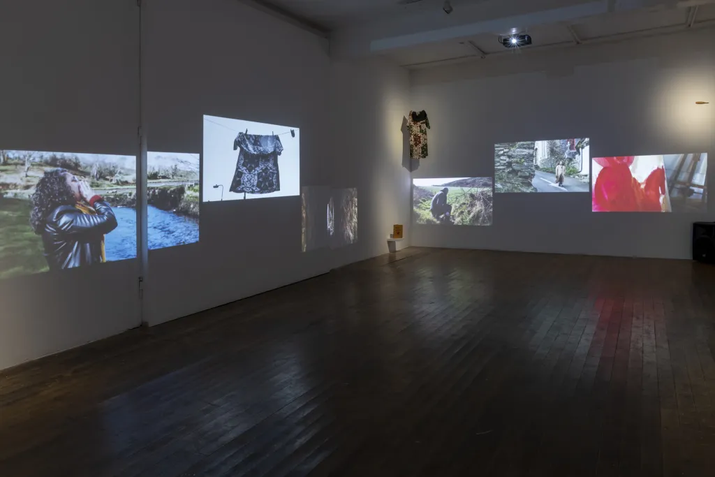 Exhibiton documentation of a white wall gallery with a wooden floor, 6 videos are projected over two walls depicitng outdoor scenes, people and clothes.