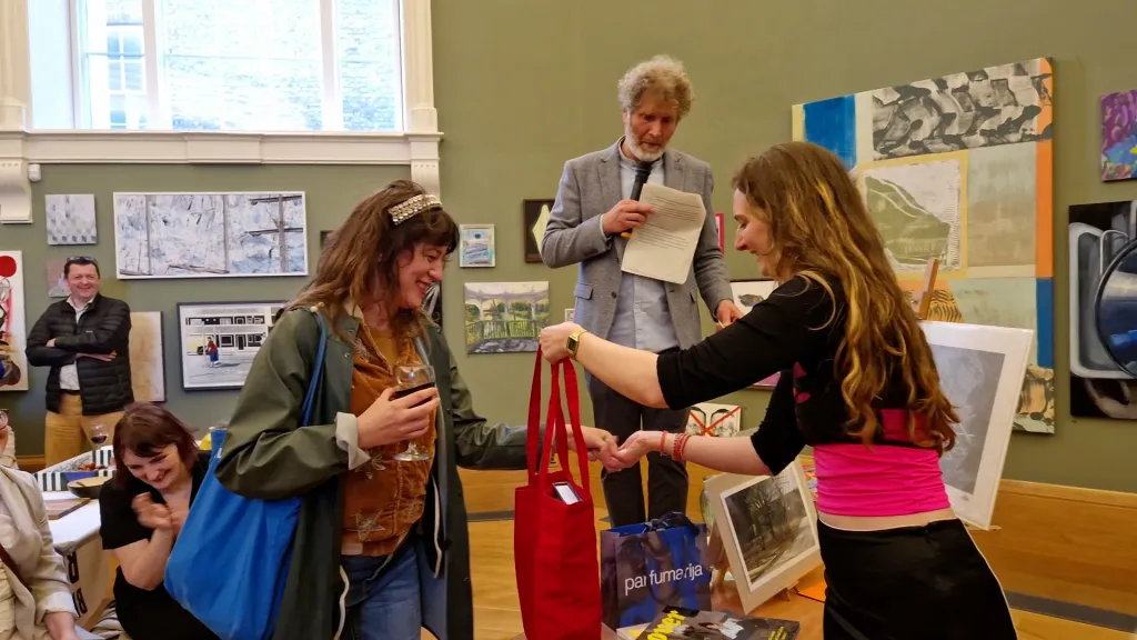 Photo of an auction, a woman hands another woman a red bag, a man holding paper stands behind them inside a room with paintings hanging all over green walls. 