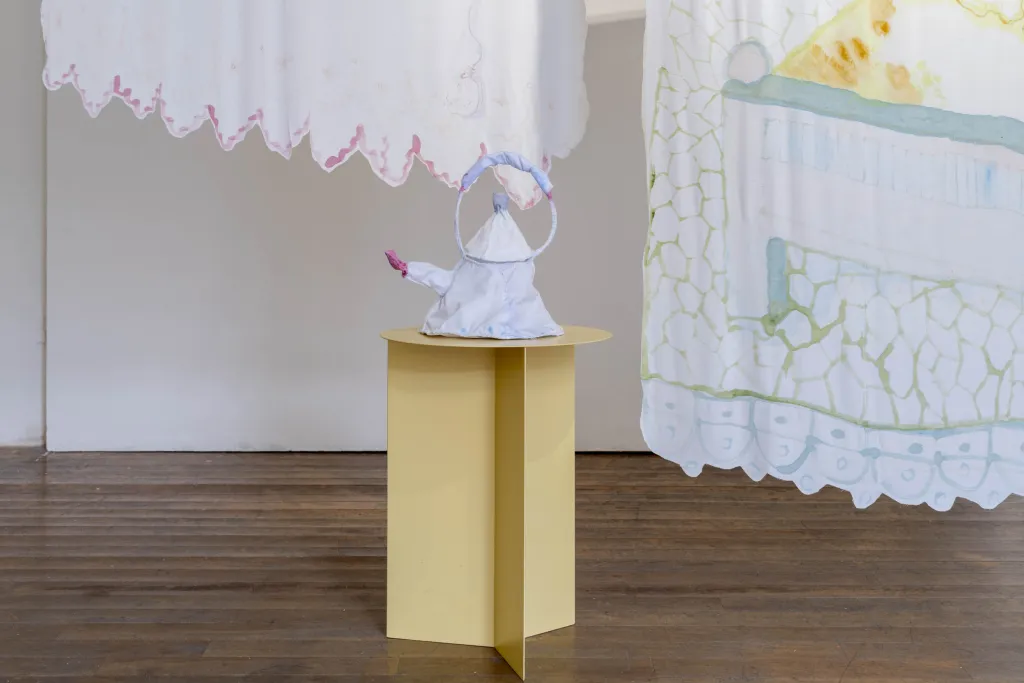 White wall gallery with wooden floor, a yellow table holds a kettle made from white cloth, two pieces of white cloth one with pink details and the other with green and yellow hang in the background.