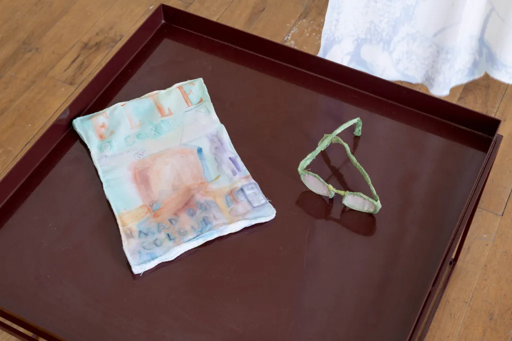 A burgundy table top holding an piece of cloth with a cover of ELLE magazine painted on top and a pair of green framed glasses made from fabric, on a wooden floor. 