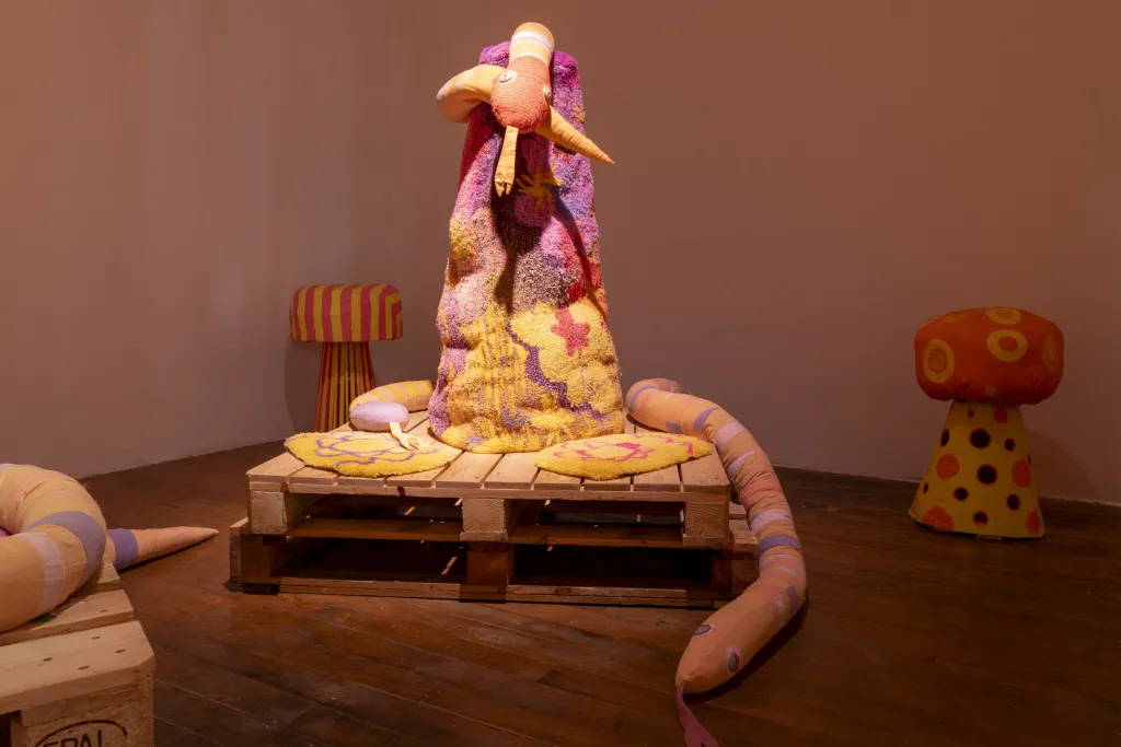 Exhibition documentation of a white wall gellery with wooden floors and low lighting, a multi-coloured fabric sculpture of a snake sits upright on two wooden palletes, two yellow and orange patterned mushroom sit in the background.