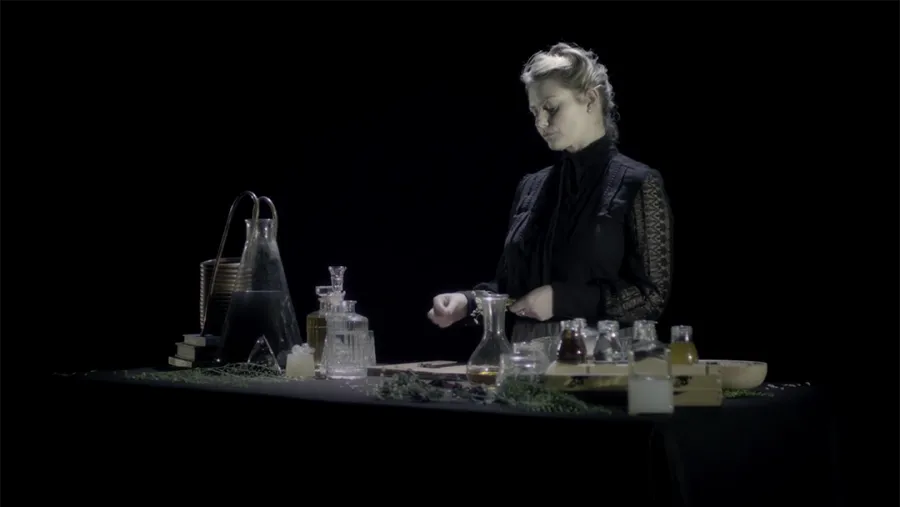 film still showing a woman dressed in black standing in front of a table with vairous sized bottle contining various liquids and plants with a black background. 