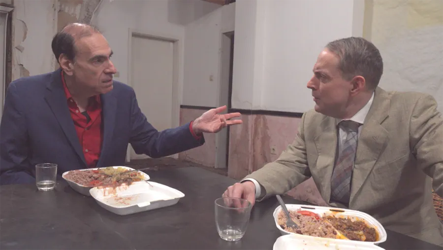 film still of two men dressed in suits having a converstaion sat at a table with take-away containers in front of them. 