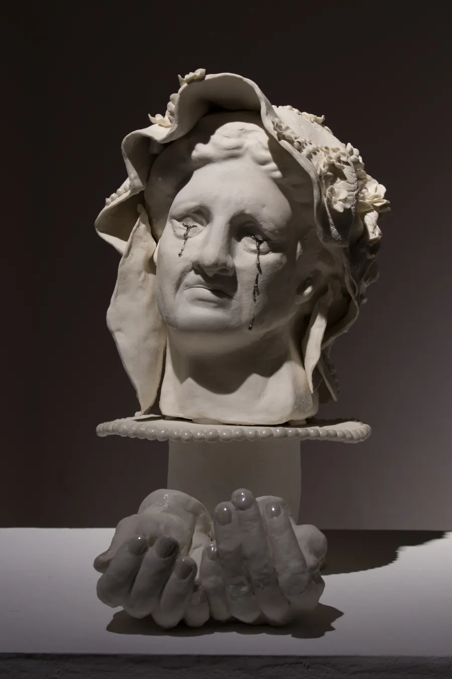 Sculpture documentation of a white bust of a womans face with a floral headdress, silver tears are rolling down the cheeks from both eyes it sits on a white ceramic cake stand and a pair of ceramic cupped hands sits in front of the bust all on a white plinth with a dark background