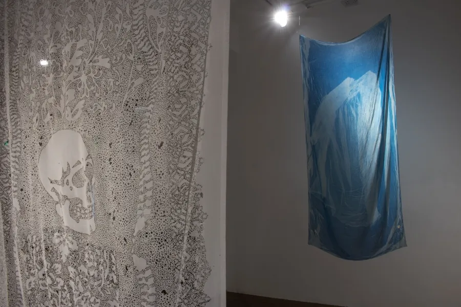 Two fabric pieces suspended from the ceiling, the left one hangs close to the camera and shows a detailed intricate pattern around a skeleton in white and grey and the second hung further from the camera on the right is a bright blue with a light blue design as if the fabric had been bleached. 