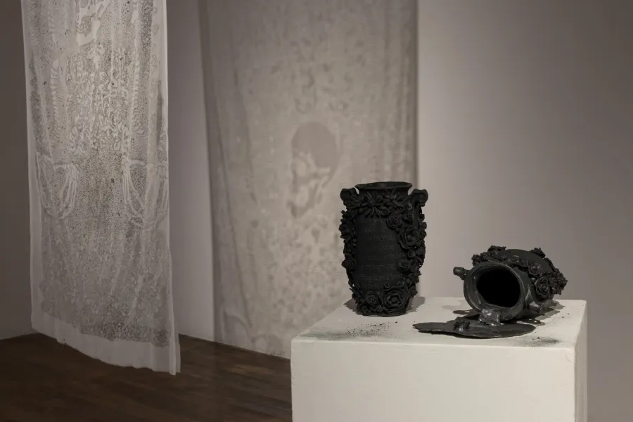 Exhibition documentation of a white wall gallery with a wooden floor, on the right the top of a white plinth holds two black ceramic vases with floral decorations around the edge of the vase, one vase sits tipped over with a black liquid spilling out on top of the plinth, to the left a white cloth with an intricate pattern is suspended from the ceiling, the light shining through it reflects on the wall behind.  