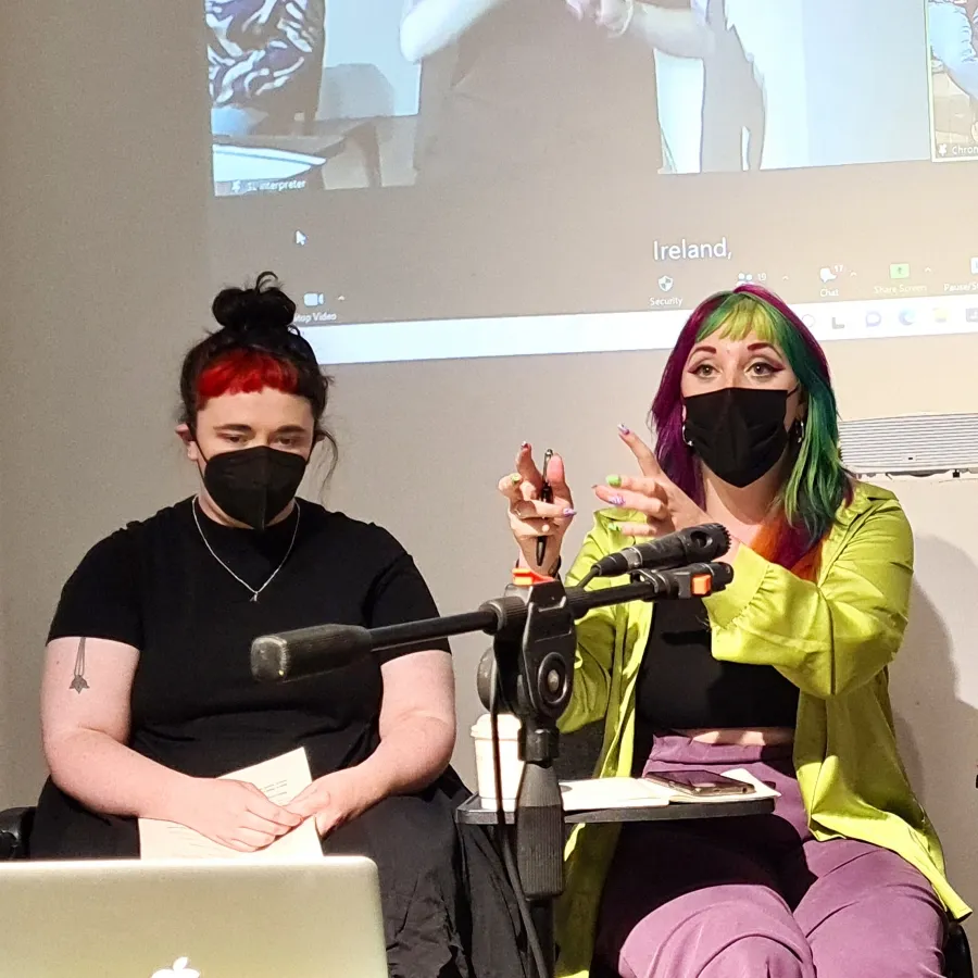 Open forum documetation two people sitting in front a projected image of a zoom call, both wear masks the person to the left sits looking at a laptop screen the person the the right is talking and describing something with their hands.