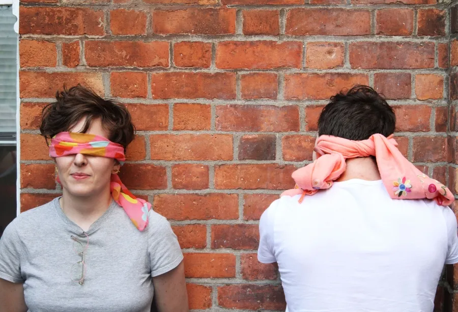 Workshop documentation of two people standing in front of a red brick wall, both wear pink fabric as blindfolds, the person to the left faces the camera and the person to the right faces the wall,