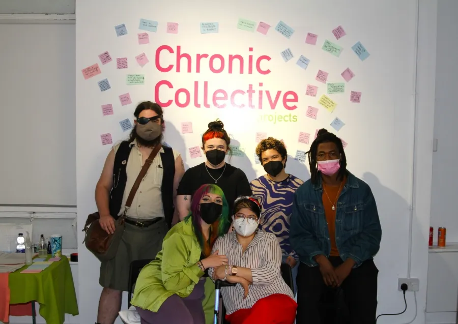 A group of people wearing face masks posing for a picture, four stand in the back, in front of them one person sits and another kneels beside them, in the background is a white wall covered in post-it notes in pastel purple, pink, green and blue surround the text 'Chronic Collective' written in a neon pink. 