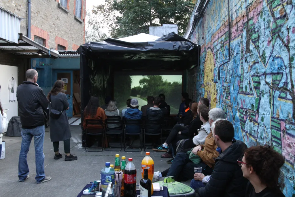 Exterier courtyard with a group of people sitting under a black gazebo watching a projection screen. 