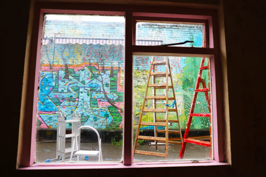 View looking outside a window with pink window frames, a red, wood, step and folded metal ladder stand in a courtyard in front of a wall painted with graffiti. 