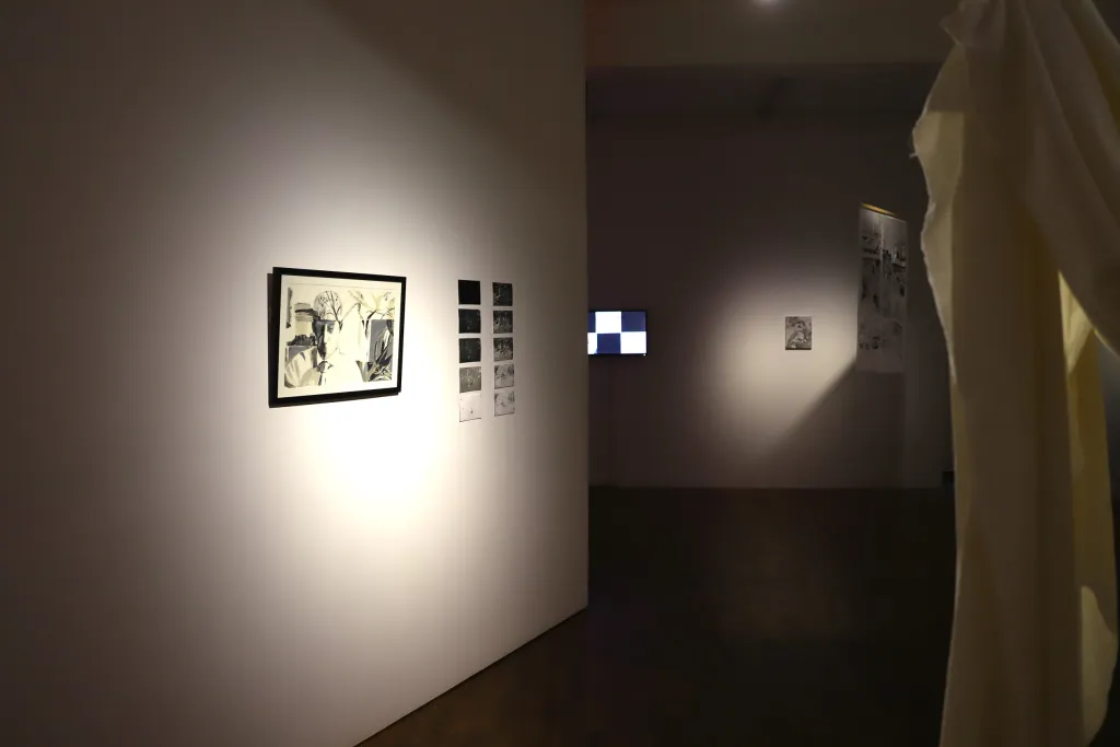 Exhibition documentation of a white wall gallery with wooden floors and low lighting, a textile piece of white fabric is drapped and hung from the ceiling, photographic prints and drawings hang on the wall, a textile piece hangs from the ceiling in the background.