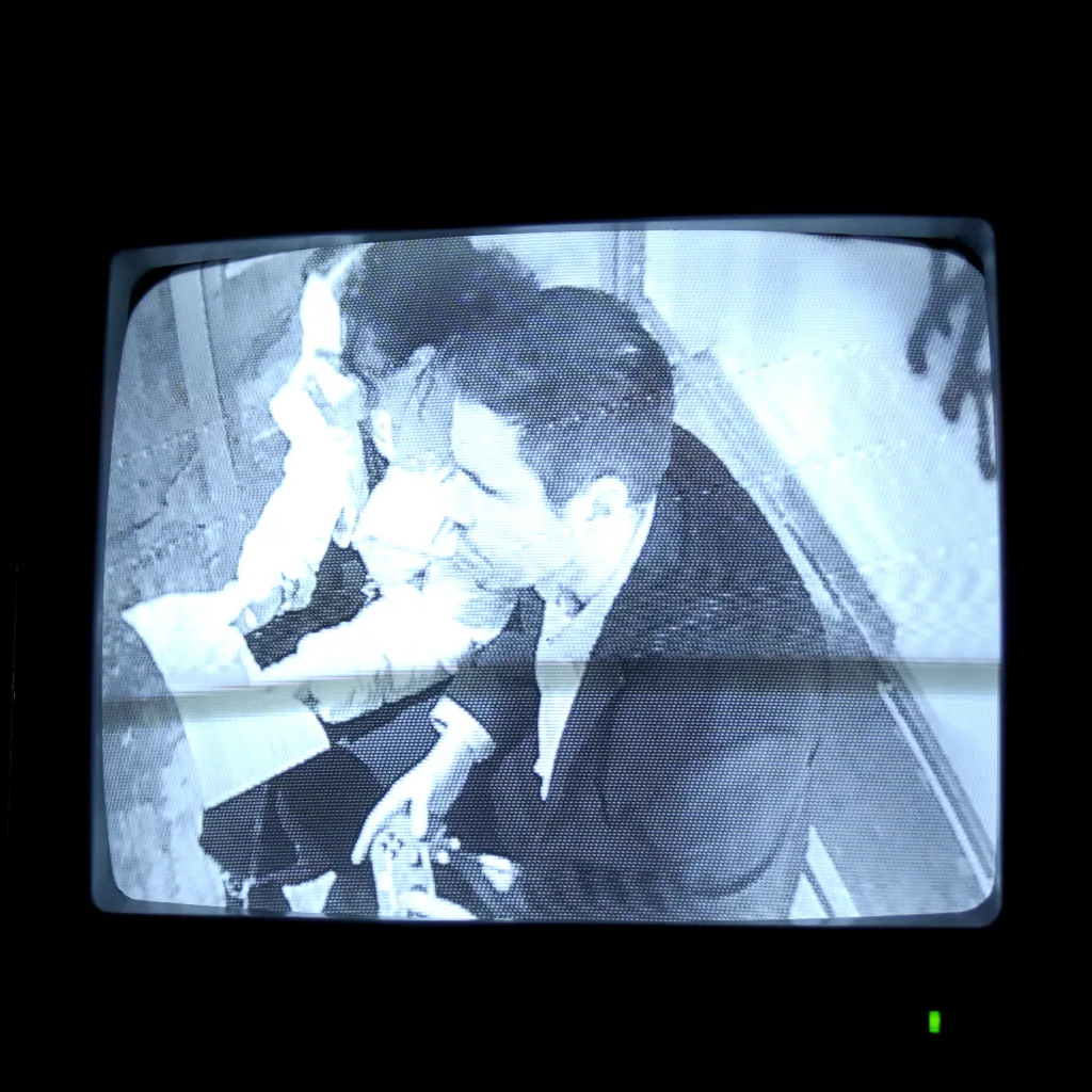 An old grey TV monitor with CCTV footage of the inside of a bus shelter with people sitting inside.