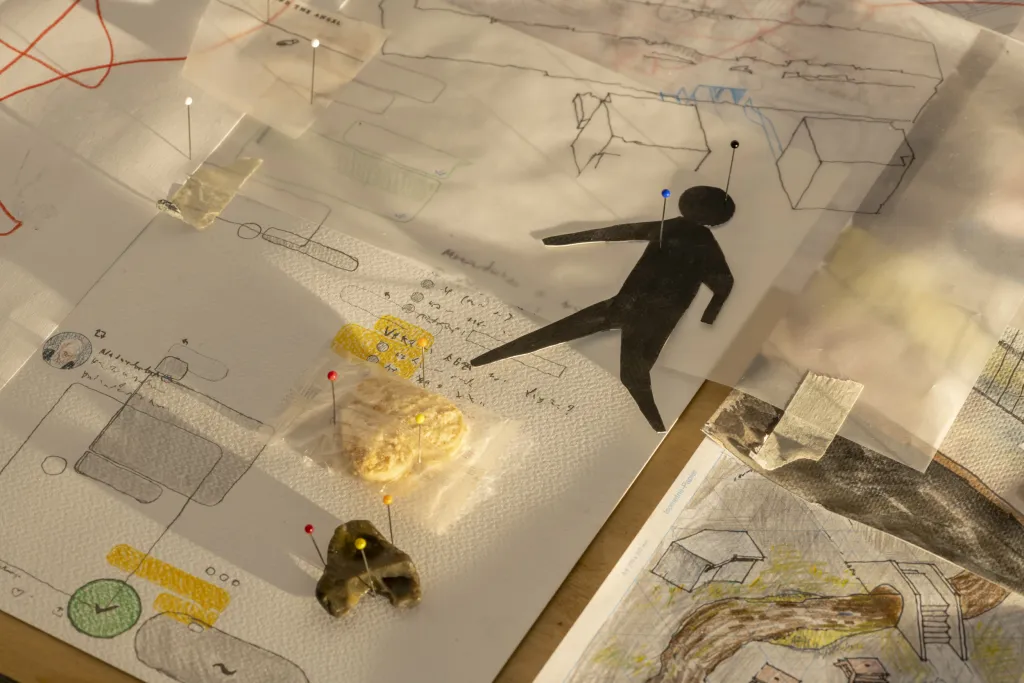 A table covered in paper with various sketches and diagrams, several pins are stuck into the paper. 