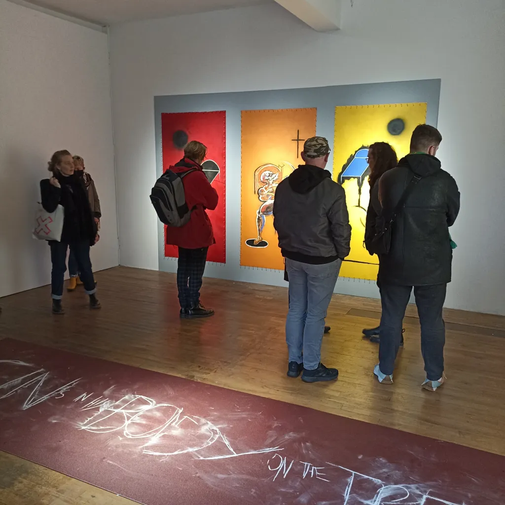 Exhibition documentation of a white wall gallery with a wooden floor, a group of 7 people stand in front of a triptych of portrait abstract paintings depicitng images of bodies and beds in vibrant red, orange and yellow, behind them on the floor a large sheet of sandpaper with scratchy text.