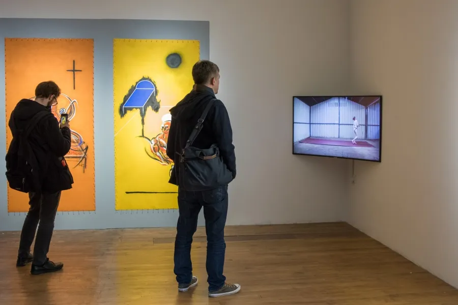 Exhibition documentation of a white wall gallery with a wooden floor, a TV hangs in the corner between 2 walls, it depicts a video of man running across a large sheet of sandpaper, on the left wall two large portrait paintings in orange and yellow  with abstract images of a bed and bodies, two men stand in front of the TV watching, the man to the left is taking a photo with his phone. 