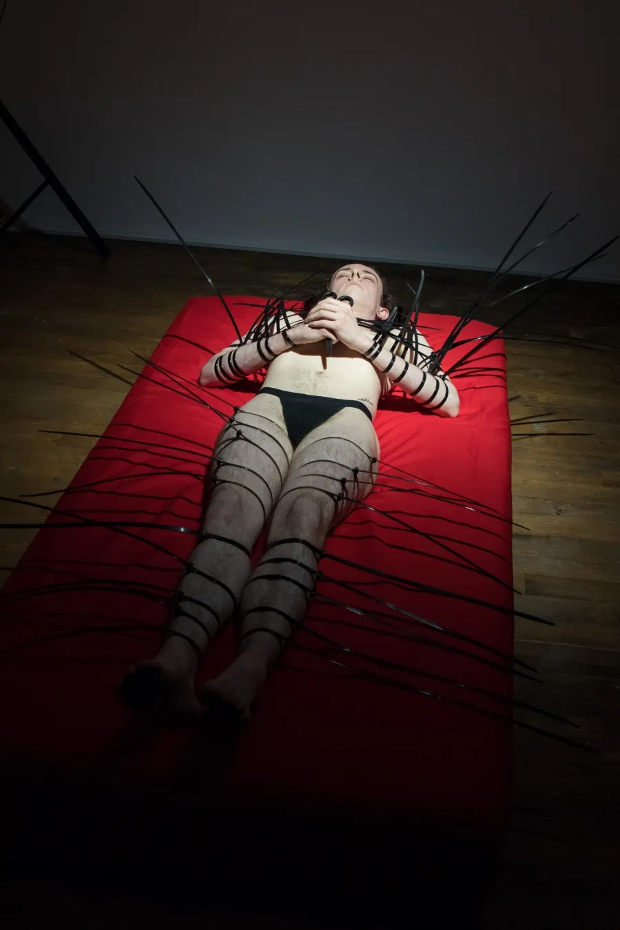 Performance documentation, a man lays in black underwear on a bright red sheet of fabric on a wooden floor, his hands hold a pair of scissors over his chest his arms and legs are tied with large black cable ties which stick out from his body, a white wall is behind him.