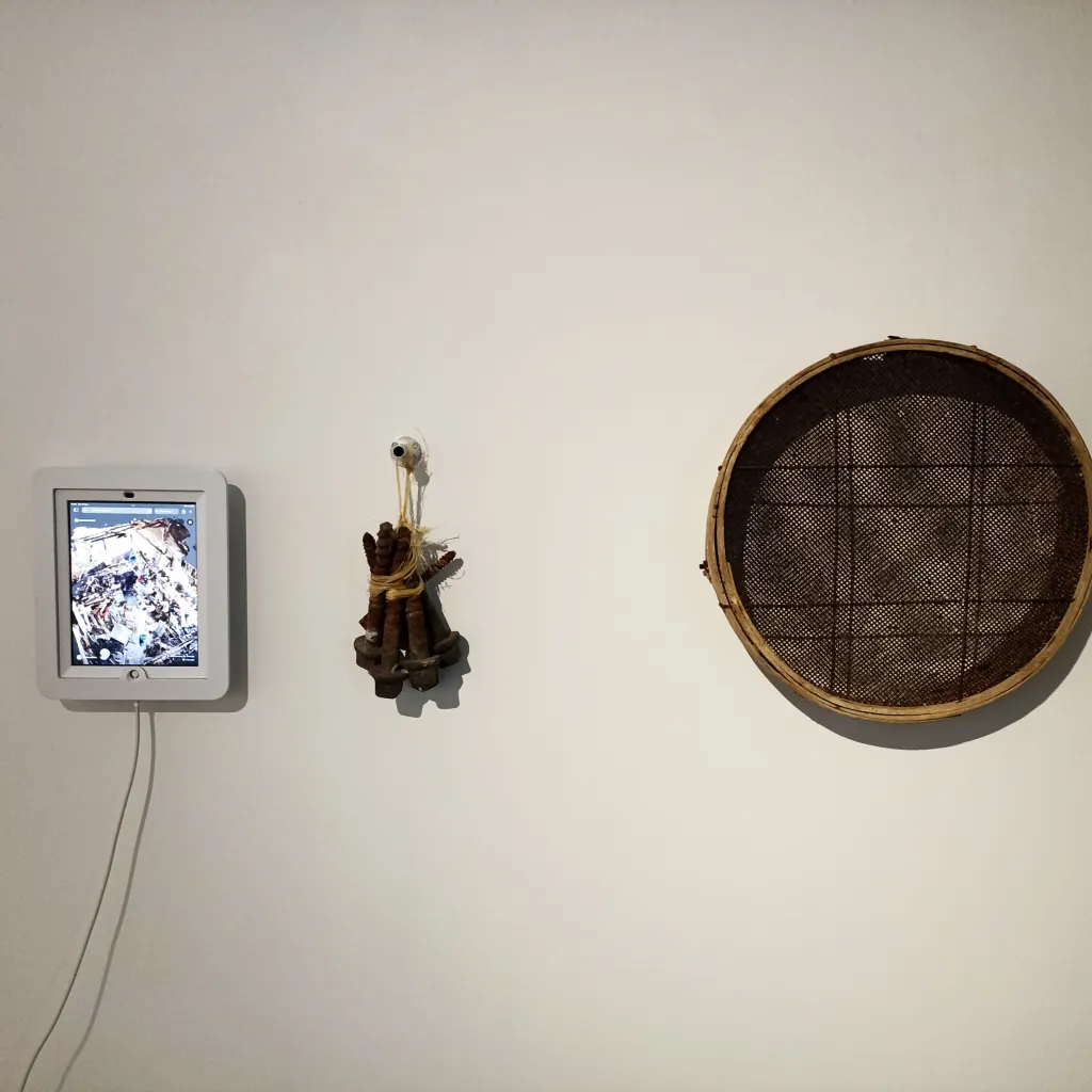 Exhibition documentation of a white gallery wall on the left s small white ipad displays a video work, in the centre a pile of large rusty metal screws tied together with twine and to the left a wooden embroidery frame with brown mesh stretched in the centre.