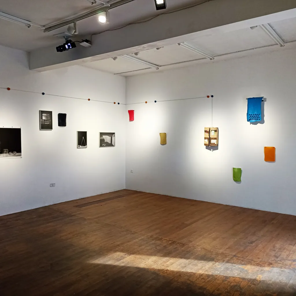 Exhibition documentation in a white wall gallery with a wooden floor, multiple prints and textile pieces hang in a various positions on the walls, in black and white and solid block colours of blue, red and green.