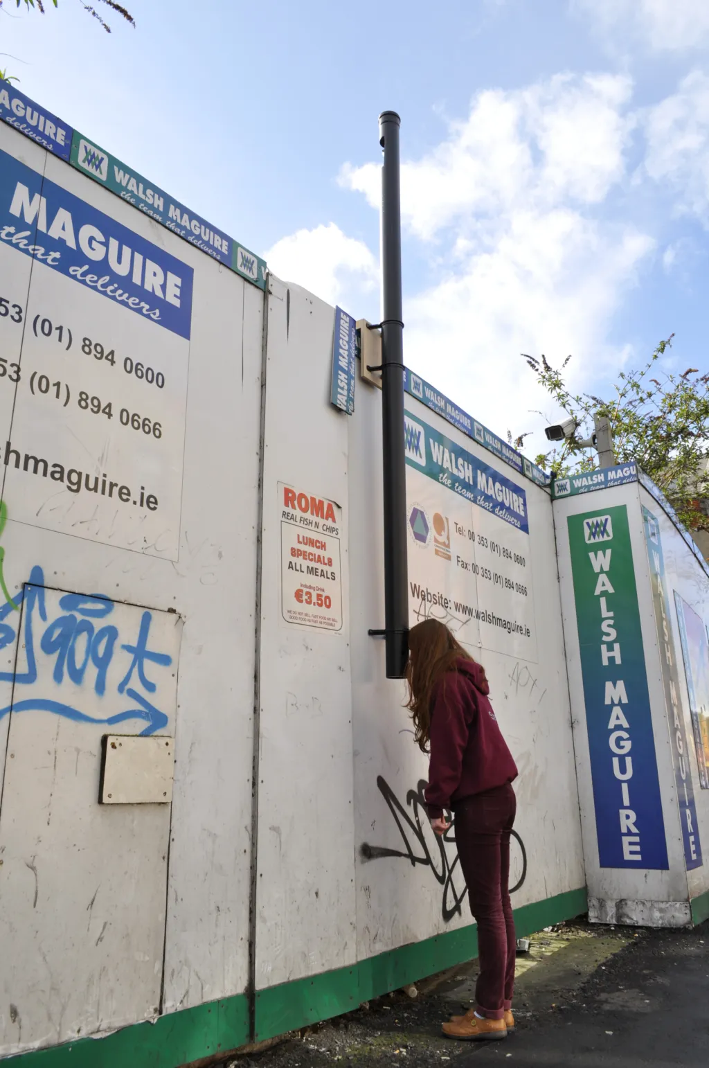 A woman peers over hoarding utilising a long metal periscope. Text on the wall reads 'Walsh Maguire, The team that delivers'