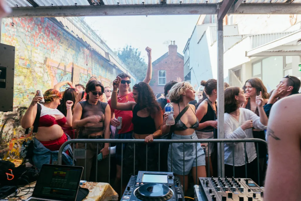 Outdoor party, people standing at a railing dancing in front of dj decks.