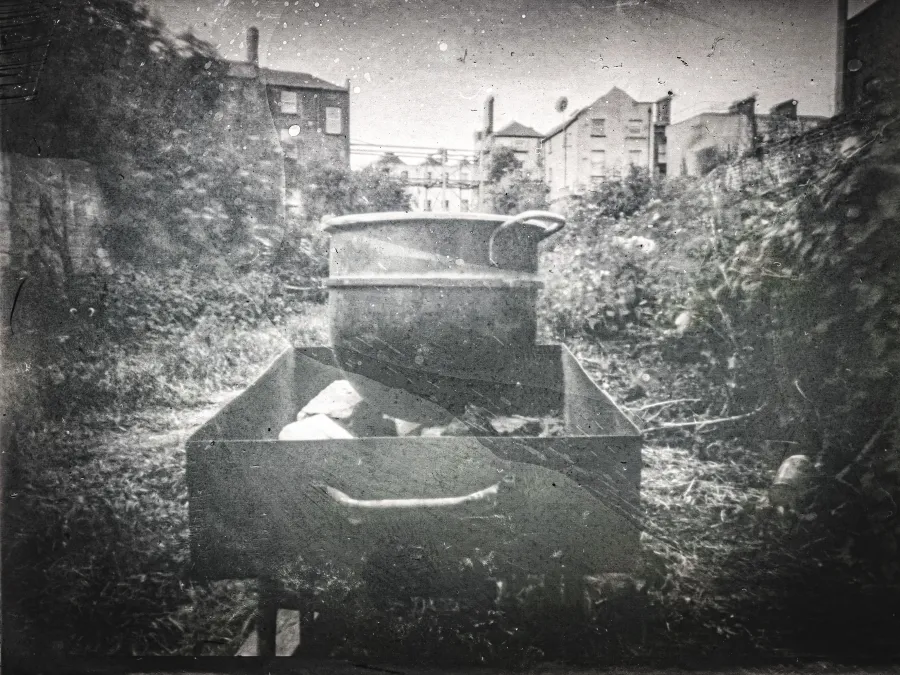 A black and white photograph showing a backgarden with houses in the background, a wooden box with pots stacked in it sits in front of the image