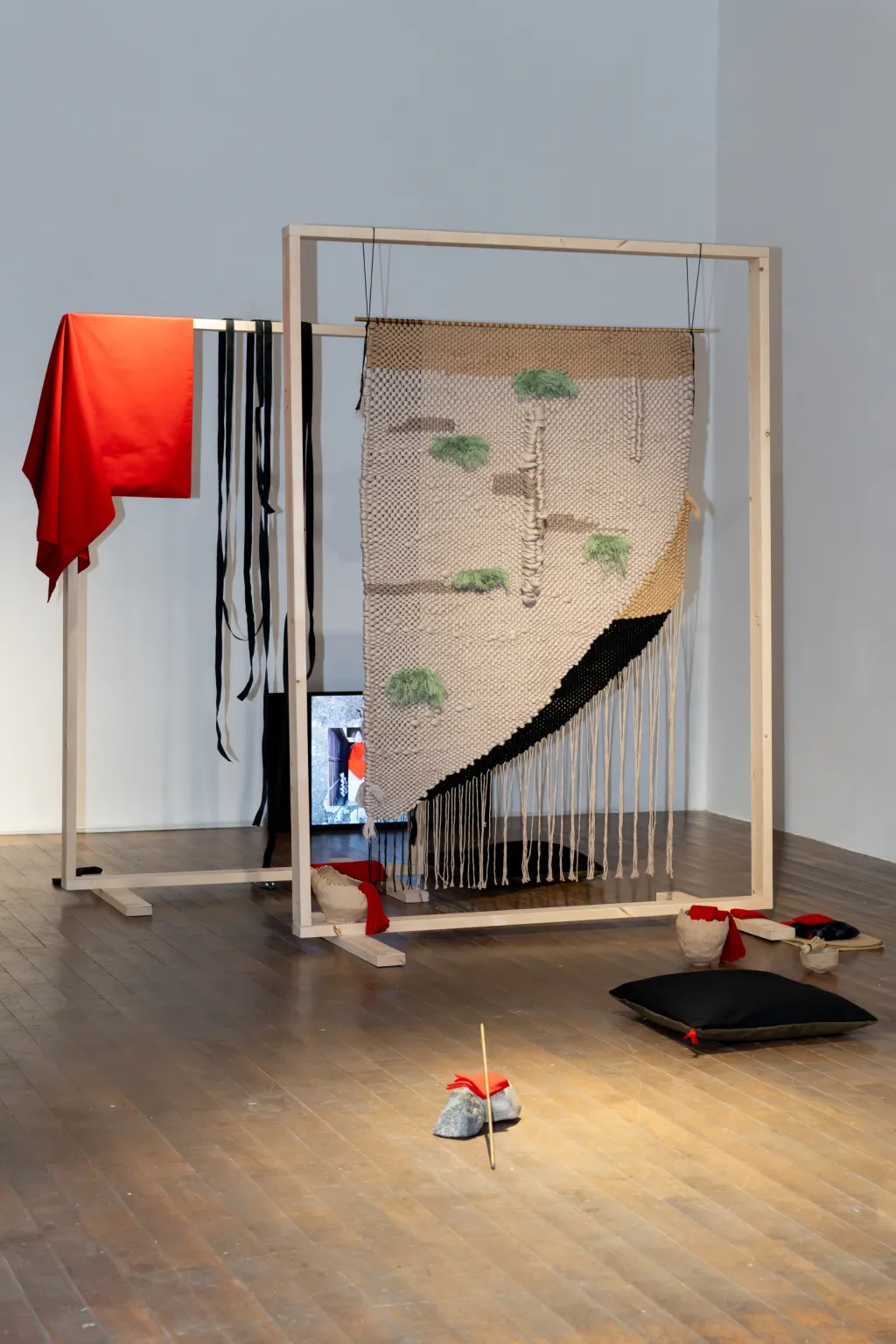 White wall gallery with wooden floor, free standing large sculpture of a wooden frame with various textiles hung in and around it