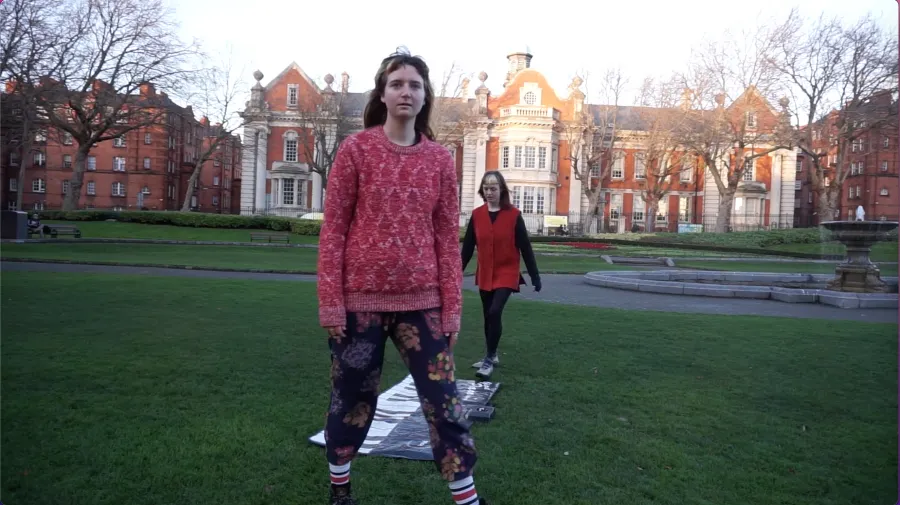 Video still depicting two women standing in a public park, the woman in the back is about to walk on a piano rug and the other woman stands on the other end facing the camera, old red brick buildings sit in the background and a fountain sits to the right.