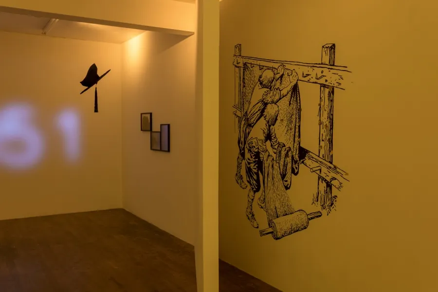 Exhibition documentation of a white wall galllery with a wooden floor, a large medievil style drawing is on the right wall, three framed images hang on the wall, a graduation cap hangs from the ceiling and the letters '51' are projected on the back wall. 