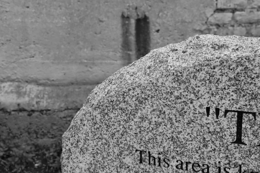 Black and white image, the corner of a granite headstone with a grey concrete wall in the background. 