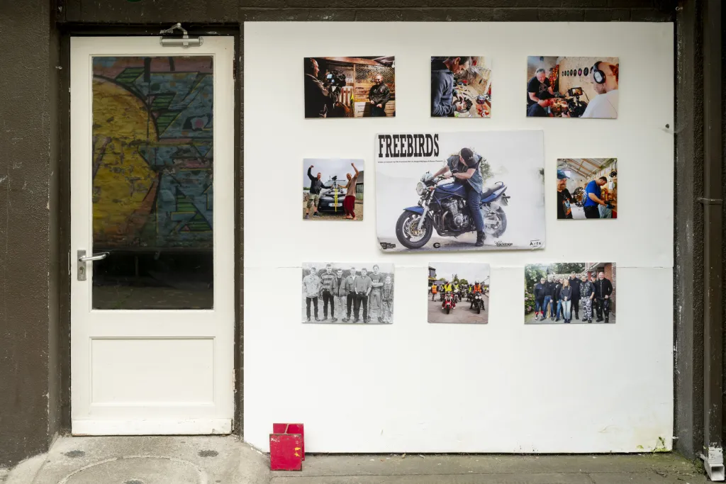 Gallery entrance from outside, a closed white door with a white wall to the right, the wall has a series of photographic prints around a large image of a man on a motercycle with 'Freebirds' written on top of the image. 