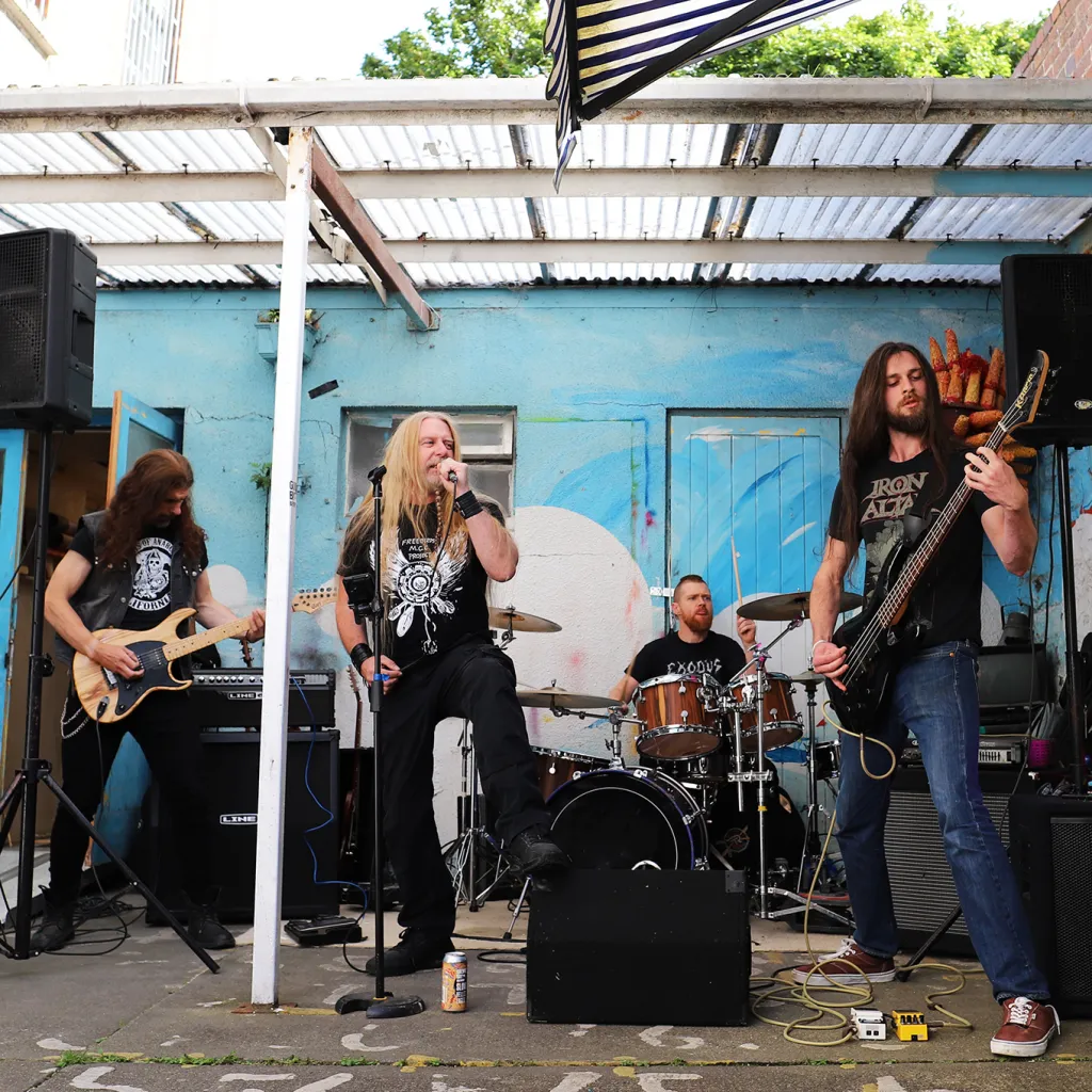 A band with two guitar players, a drummer and a singer playing outside under a shelter with a blue wall behind them.