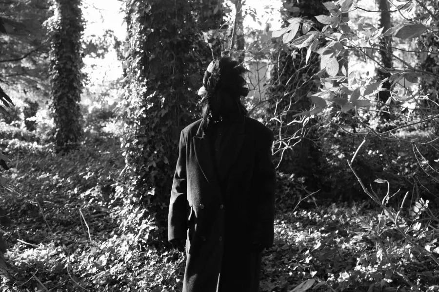 Black and white video still, figure in forest wearing a suit with a feathered mask