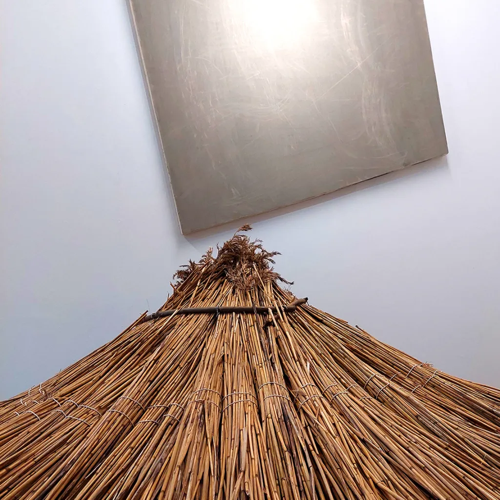 The top of a pyramid made from dried bamboo and a scratched grey painting hangs diagonally on a white wall in the background.