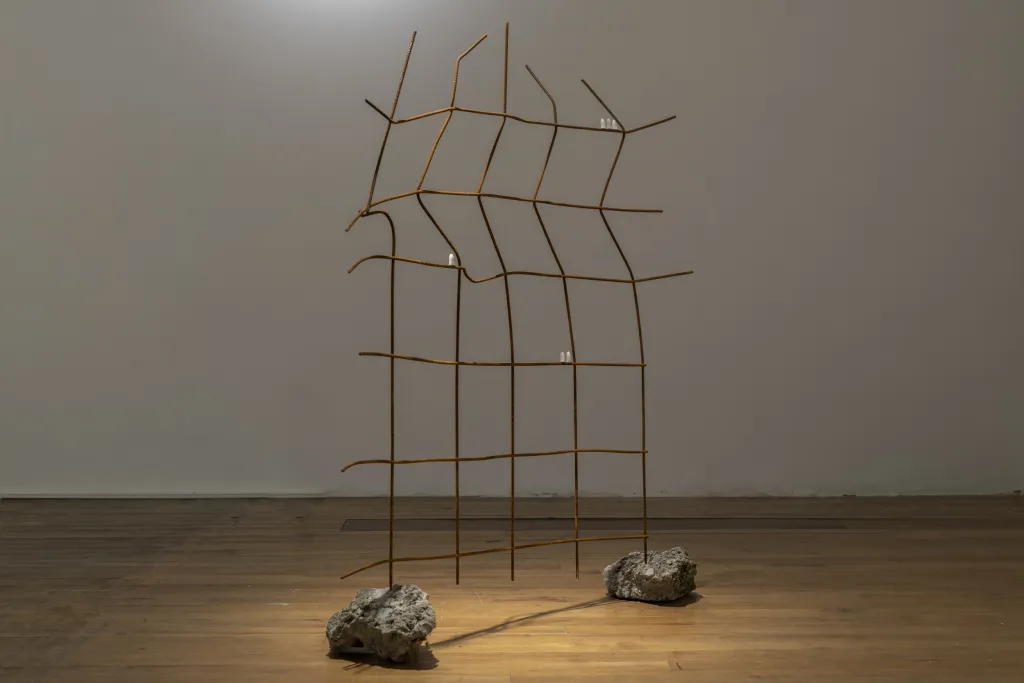 White wall gallery with a wooden floor, a sculpture made from six horizontal rods and 5 vertcal rods bent slightly out of shape, the two end rods are held up by two large grey rocks.