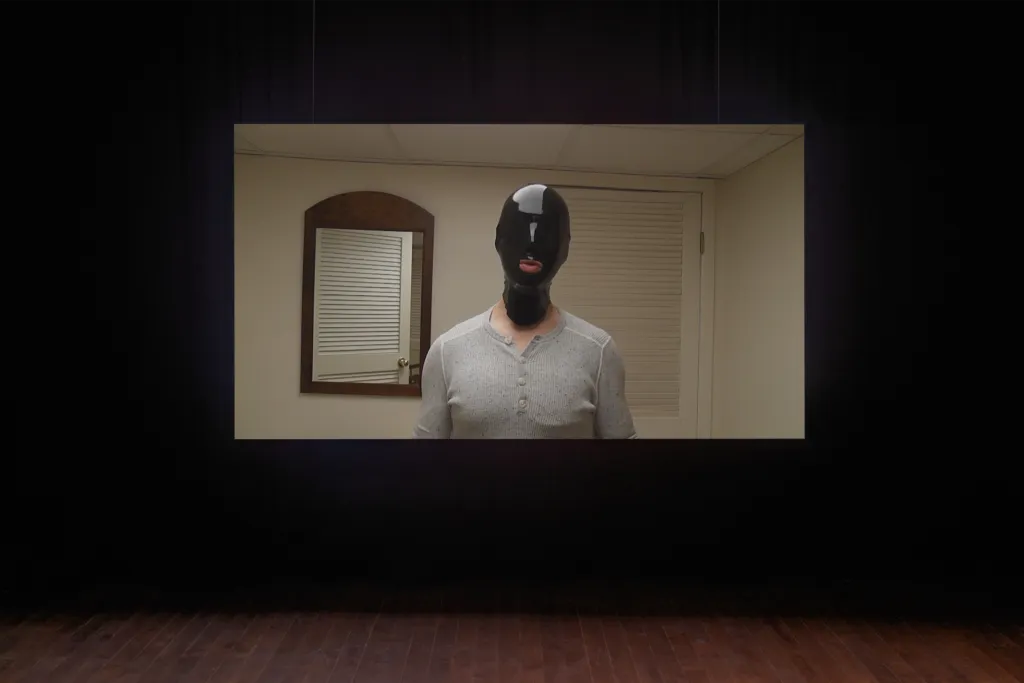 An image of a man wearing a full face black latex mask projected on a suspended screen, black curtains in background and wooden floor at the bottom of the photo.