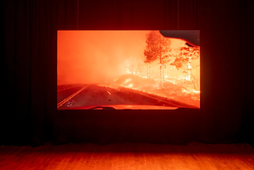 An image of a wildfire through a cars front window projected on a suspended screen, black curtains hang in the background , a wooden floor is at the bottom of the image.