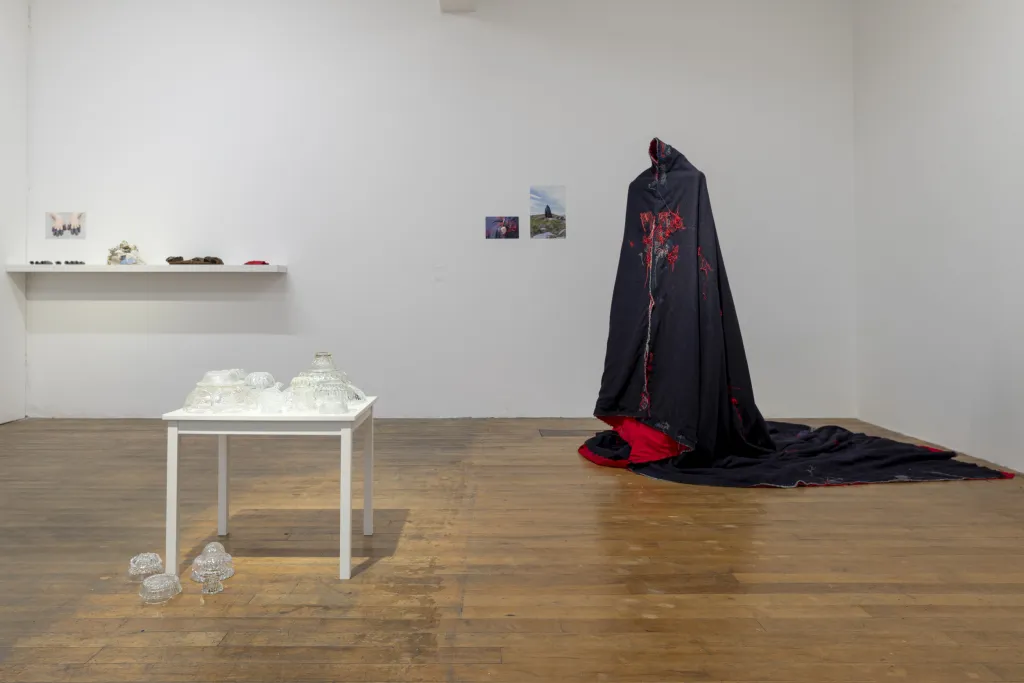 Exhibition documentation, white walled gallery wooden floor, a white table with glass bowls of various sizes to the left of the image and a navy cloak with red details is to the right.