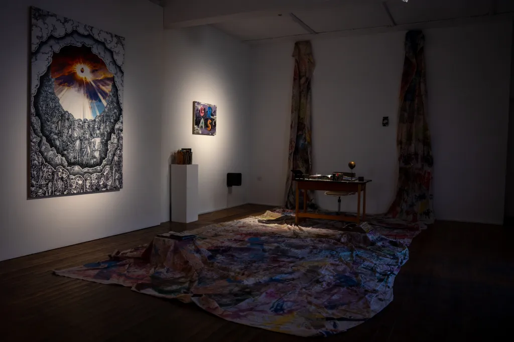 exhibition installation, a desk in the left of the room sits on a tarp covered in paint hanging from the wall, two paintings hang on a white wall to the left, a white plinth with books of various sizes sits againts the wall with low lighting.