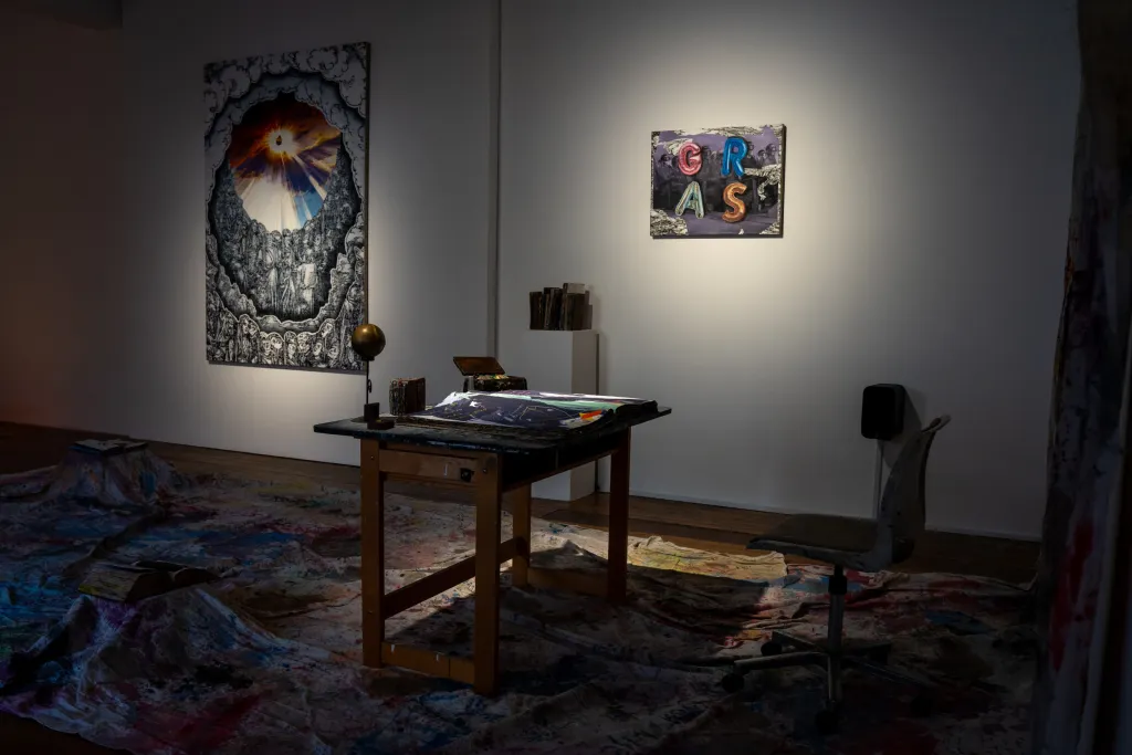 exhibition installation, a desk in the center of the room sits on a tarp covered in paint hanging from the right wall, two paintings hang on a white wall in the background with low lighting.