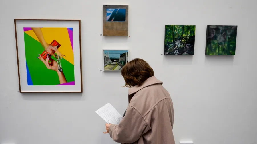 whhite wall with various sized artworks hung on it, a woman reading an infirmation sheet stands in front of the wall.