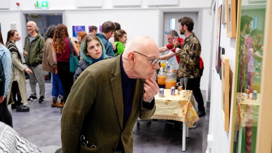 Group of people at an exhibition opening, a man leaned over looking at a painting on the wall in the forefront.