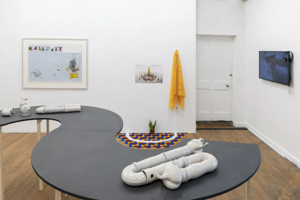 Exhibition documentation of a white wall gallery with wooden floor, a sculpture stands on the floor made from cast cement ressembling a gas tank, with a detail of a face with its tongue out, yellow tubing and a metal trangle on the floor.