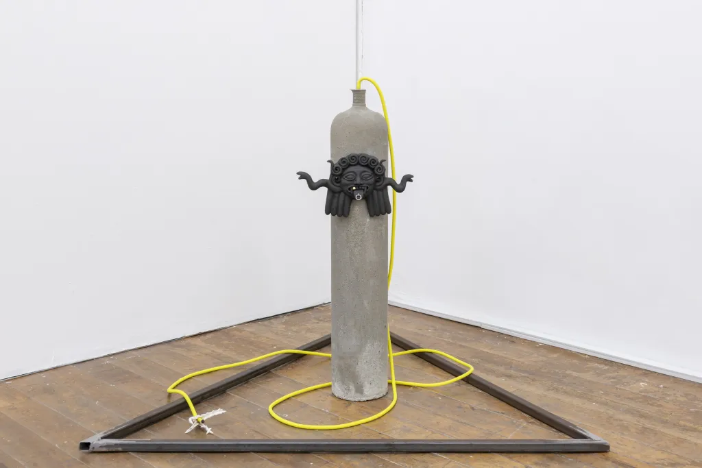 Exhibition documentation of a white wall gallery with wooden floor, a sculpture stands on the floor made from cast cement ressembling a gas tank, with a detail of a face with its tongue out, yellow tubing and a metal trangle on the floor. 