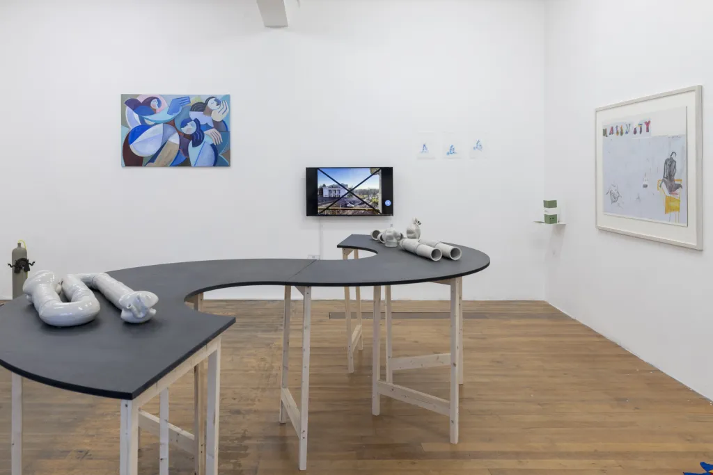Exhibition documentation of a white wall gallery with wooden floors, a curving grey table top holds a white sculptures resembling pipes, a TV displaying video work, and two paintings hang on the wall.