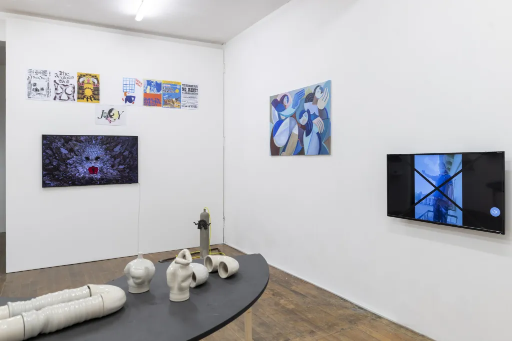 Exhibition documentation of a white wall gallery with wooden floors, a curving grey table top holds a white sculptures resembling pipes, 2 TVs displaying video works, paintings and drawings hang on the wall.