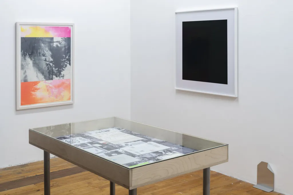 white wall gallery with wooden floor, a table with glass top displaying prints in the middle of two framed works hung on the left and right. 